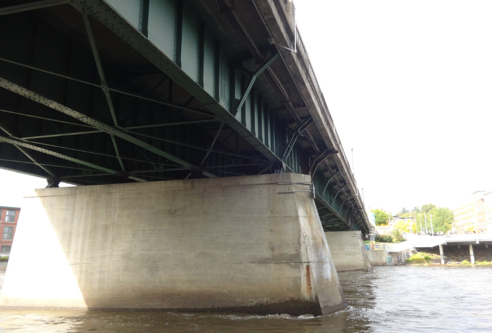 A concrete and steel bridge over a body of water, with the underside of the bridge and its support pillars visible. Buildings are faintly visible in the background under a clear sky.