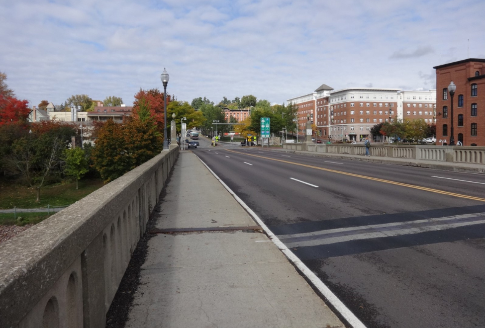 A wide city street with multiple lanes, bordered by a clean sidewalk and a concrete barrier. Buildings and trees line the street, and street lamps are installed along the sidewalk. The partly cloudy sky adds to the urban scene.