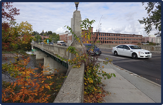 A concrete bridge with a stone pillar, with vehicles driving across the bridge, surrounded by trees with changing leaves, and buildings in the background under a partly cloudy sky.