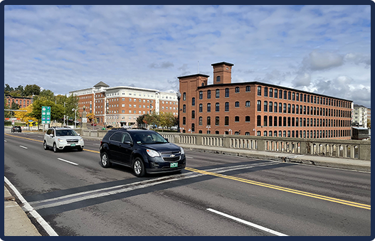 A cityscape with two cars on a bridge, surrounded by large brick buildings under a partly cloudy sky.