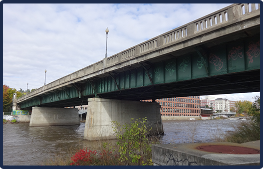 A concrete bridge with green steel underside spans a river, with buildings and trees visible in the background under a partly cloudy sky.